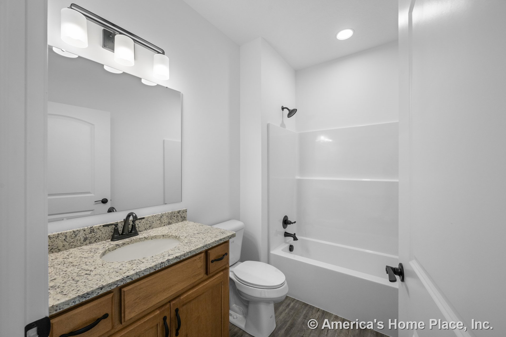 Single vanity with granite countertop and wood cabinetry, tub-shower combination with white surround, wall-mounted mirror above vanity, three-light fixture, wood-look flooring