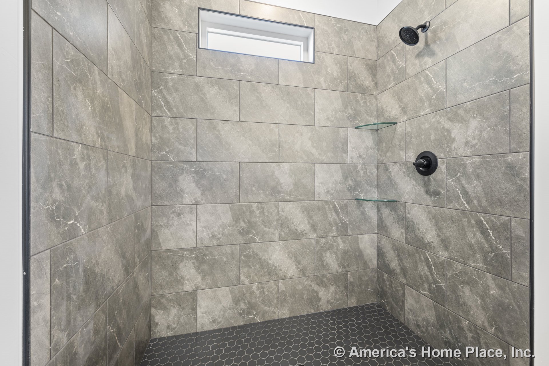 Walk-in shower with large gray stone-look wall tiles, black hexagonal floor tile, built-in glass corner shelves, matte black shower fixtures, and a horizontal window.