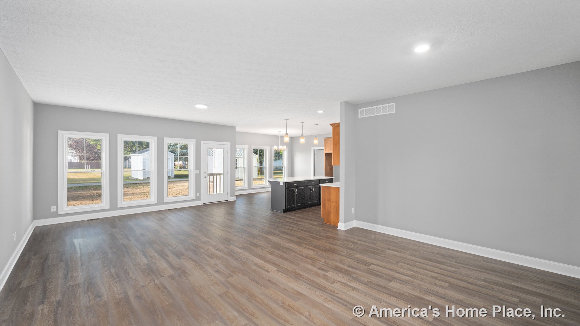 Open concept living area with four large windows, glass patio door, vinyl plank flooring, white trim and baseboards, kitchen island featuring dark cabinetry, pendant lights above