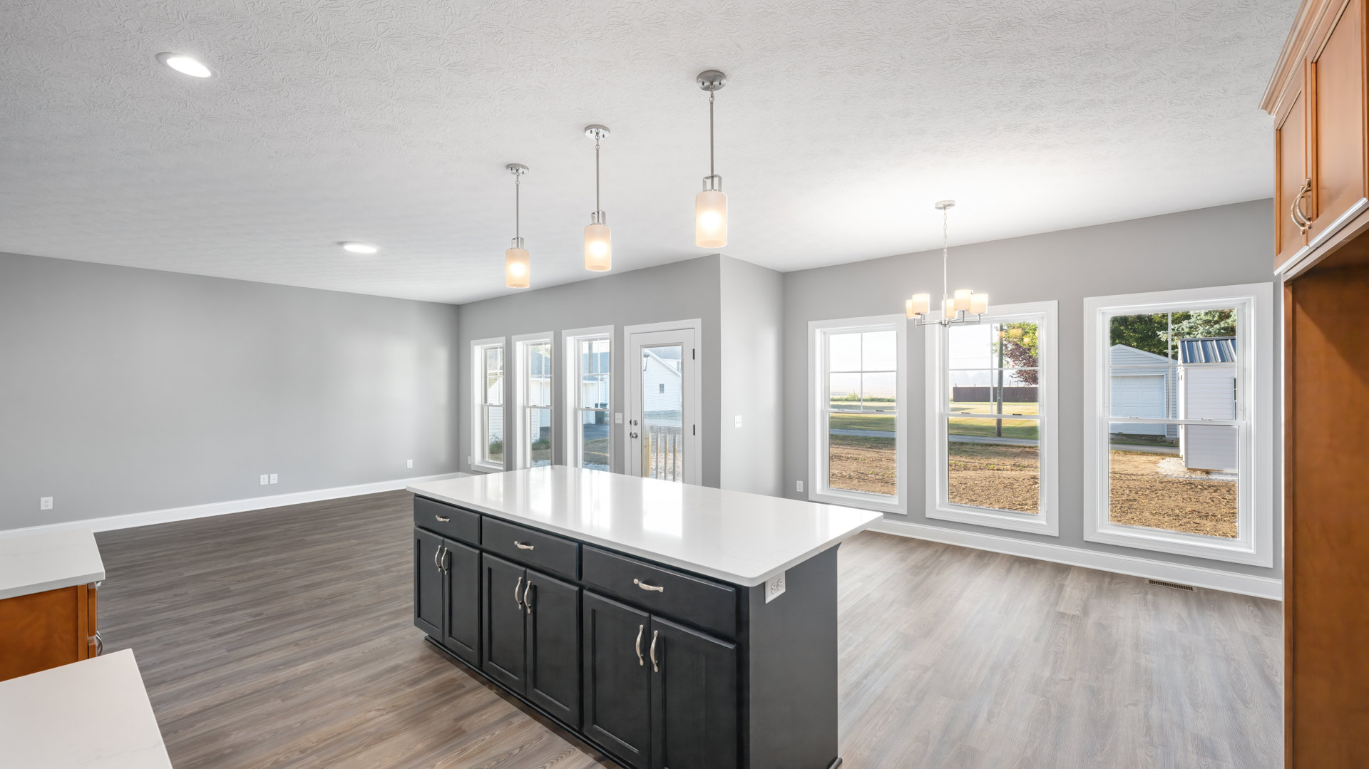Spacious kitchen featuring a large island with black cabinets, white countertop, pendant lighting, wide windows overlooking a yard, light wood flooring, and modern cabinetry.
