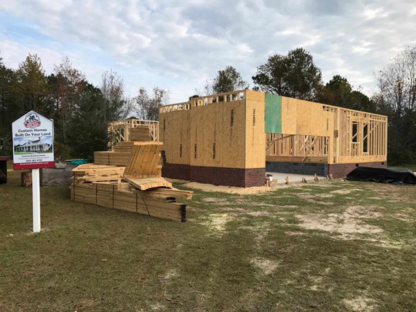 Framed house under construction with exposed wood beams, stacked lumber in foreground, grassy lot, cloudy sky, and surrounding trees