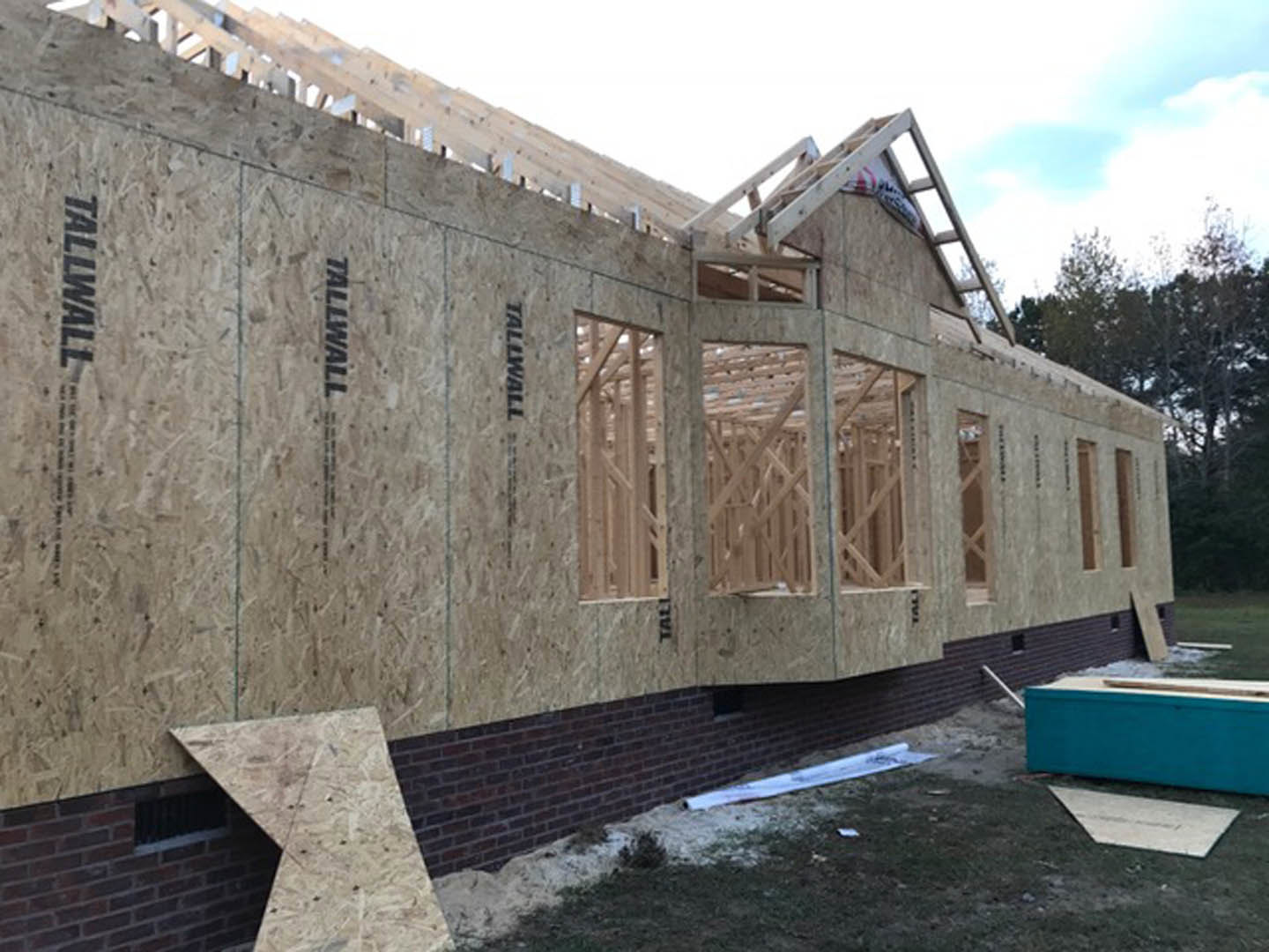 Partially built brick exterior wall with exposed concrete foundation, blue utility box, and unfinished roof under cloudy sky