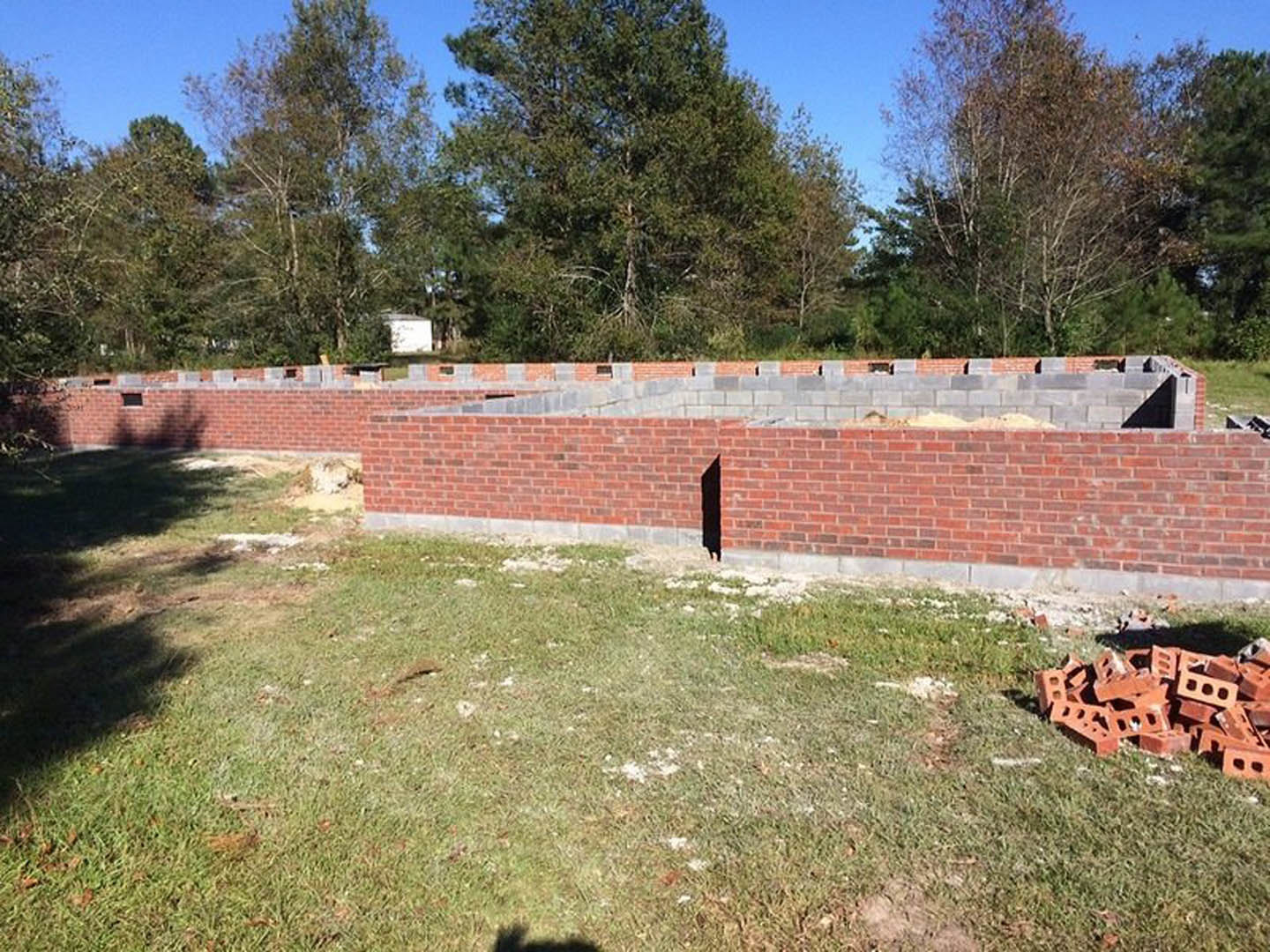 Red brick wall with a large hole, scattered bricks on grassy ground, leafy tree casting shadow, group of trees in background, blue sky overhead