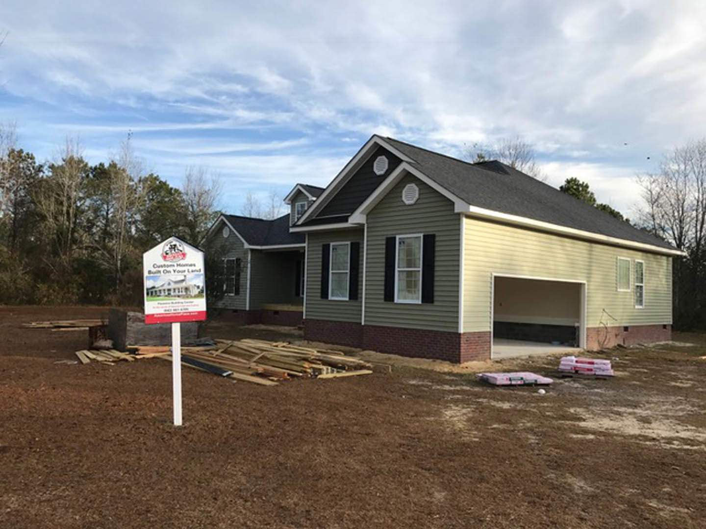 Two-story home under construction with exposed framing, attached garage, white-framed windows, and a real estate sign featuring a house illustration on the front lawn