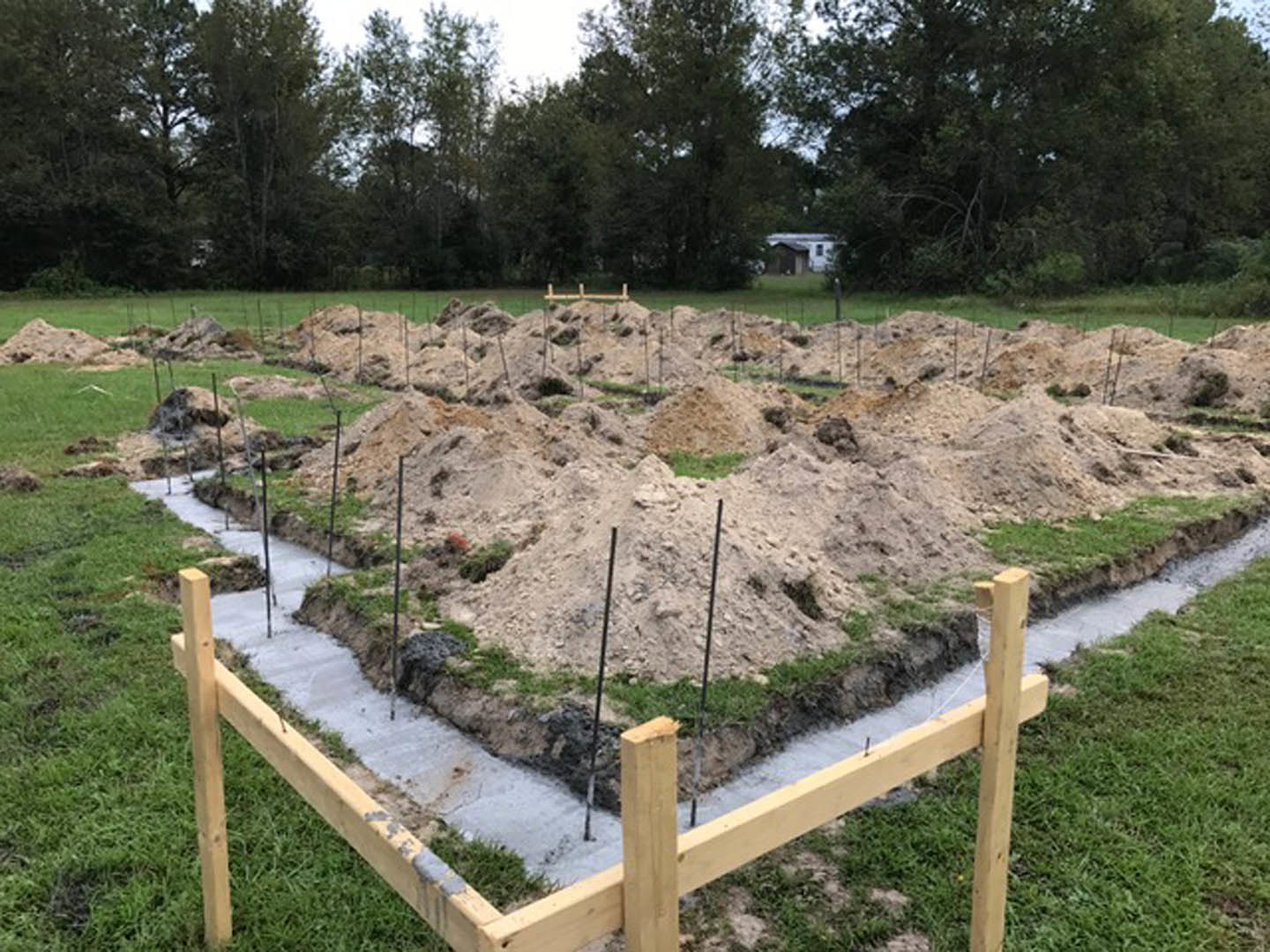 Fenced construction site with dirt, grass, and scattered poles; trees in the background; pile of dirt and rocks in foreground; partially visible house.