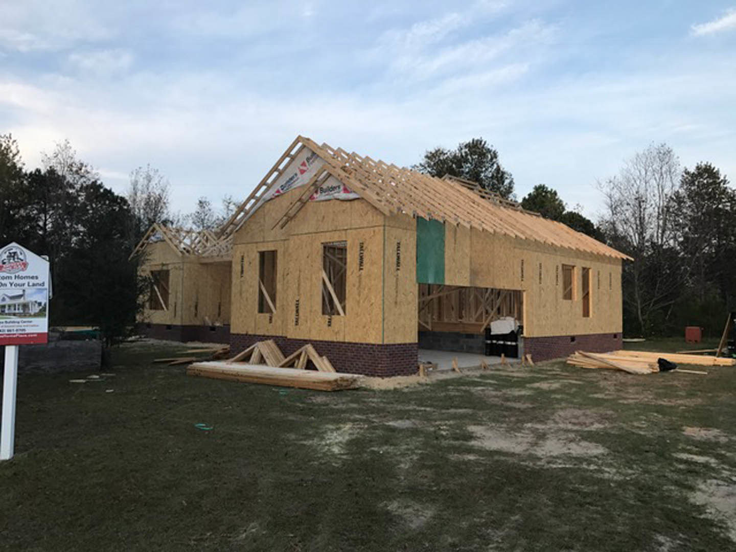Partially built house framed with exposed wood beams, surrounded by grass and mature trees under a cloudy sky