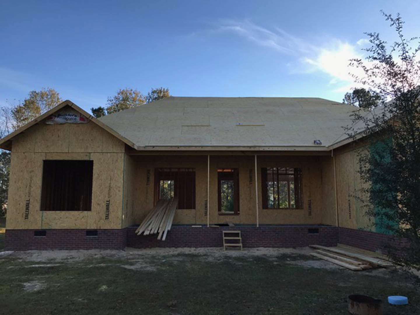 Partially built home exterior with exposed brick walls, black door framed in white, barred window, wood planks stacked on porch, and construction materials scattered in front.