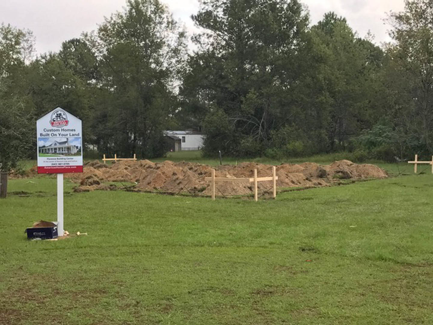 Wooden sign with white text stands in grassy field bordered by trees under blue sky