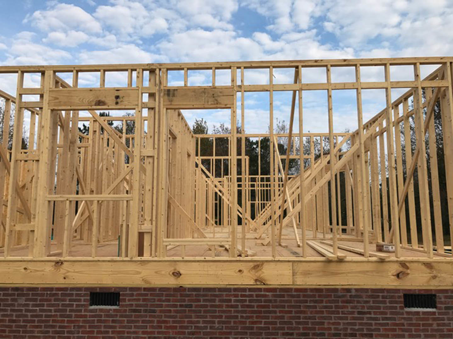 Wood-framed house under construction with exposed beams, partial brick wall, and blue sky with scattered clouds in the background