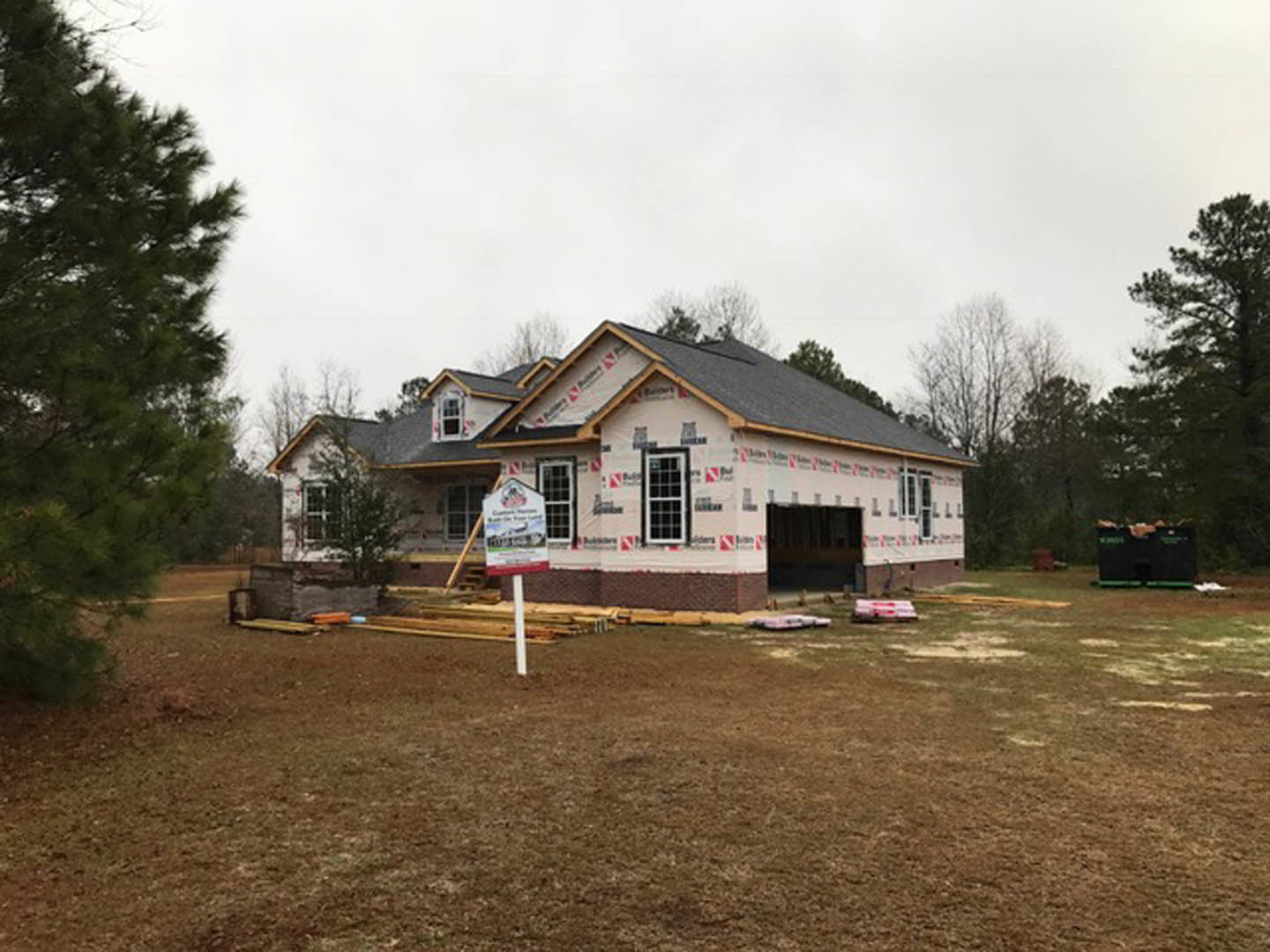 Framed custom home under construction on a dirt lot, surrounded by mature trees and cloudy sky, with a construction sign in the foreground