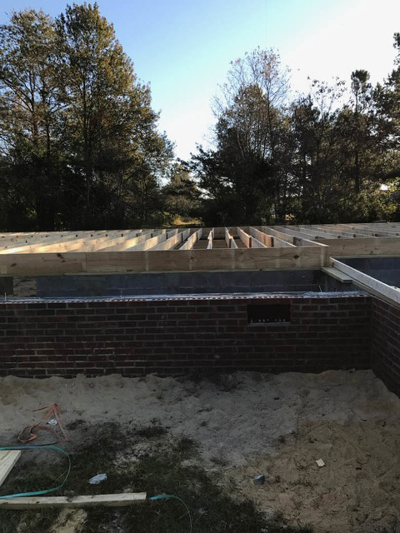 Partially built brick exterior wall with concrete foundation, person standing on wall, snow covering ground, mature trees and blue sky in background