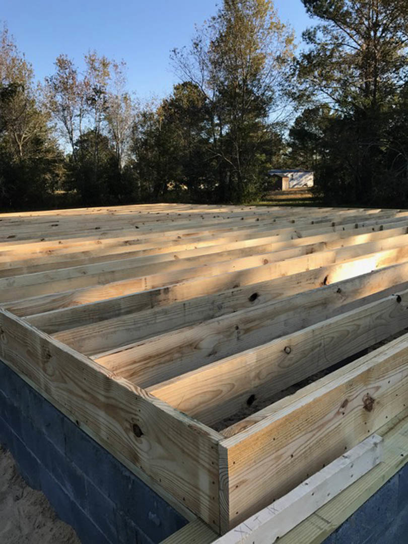 Unfinished wooden framing with exposed beams and planks on a deck, surrounded by trees under a clear sky