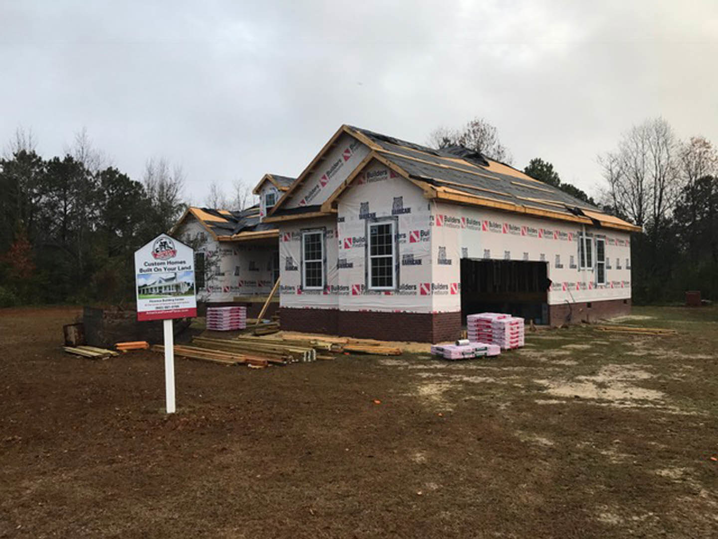 Partially built home with shingled roof, multi-pane windows, and construction sign posted in front; boxes stacked near foundation, surrounded by dirt and scattered trees under