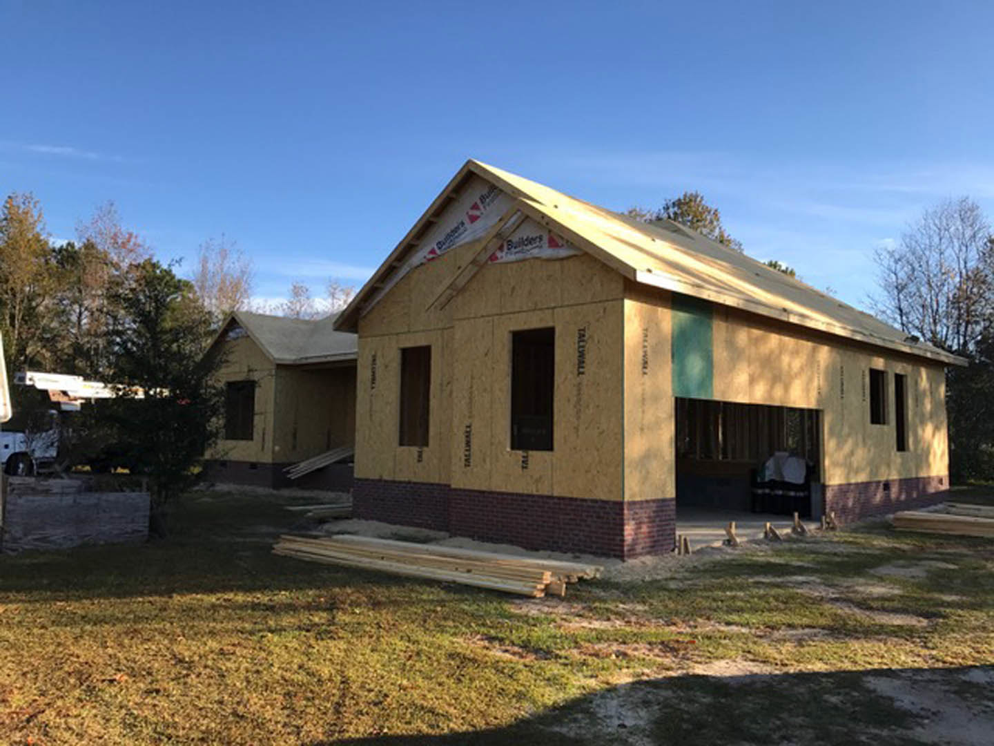 Two-story house under construction with exposed framing, partially finished roof, and light-colored siding, surrounded by grassy yard and mature trees under a clear sky