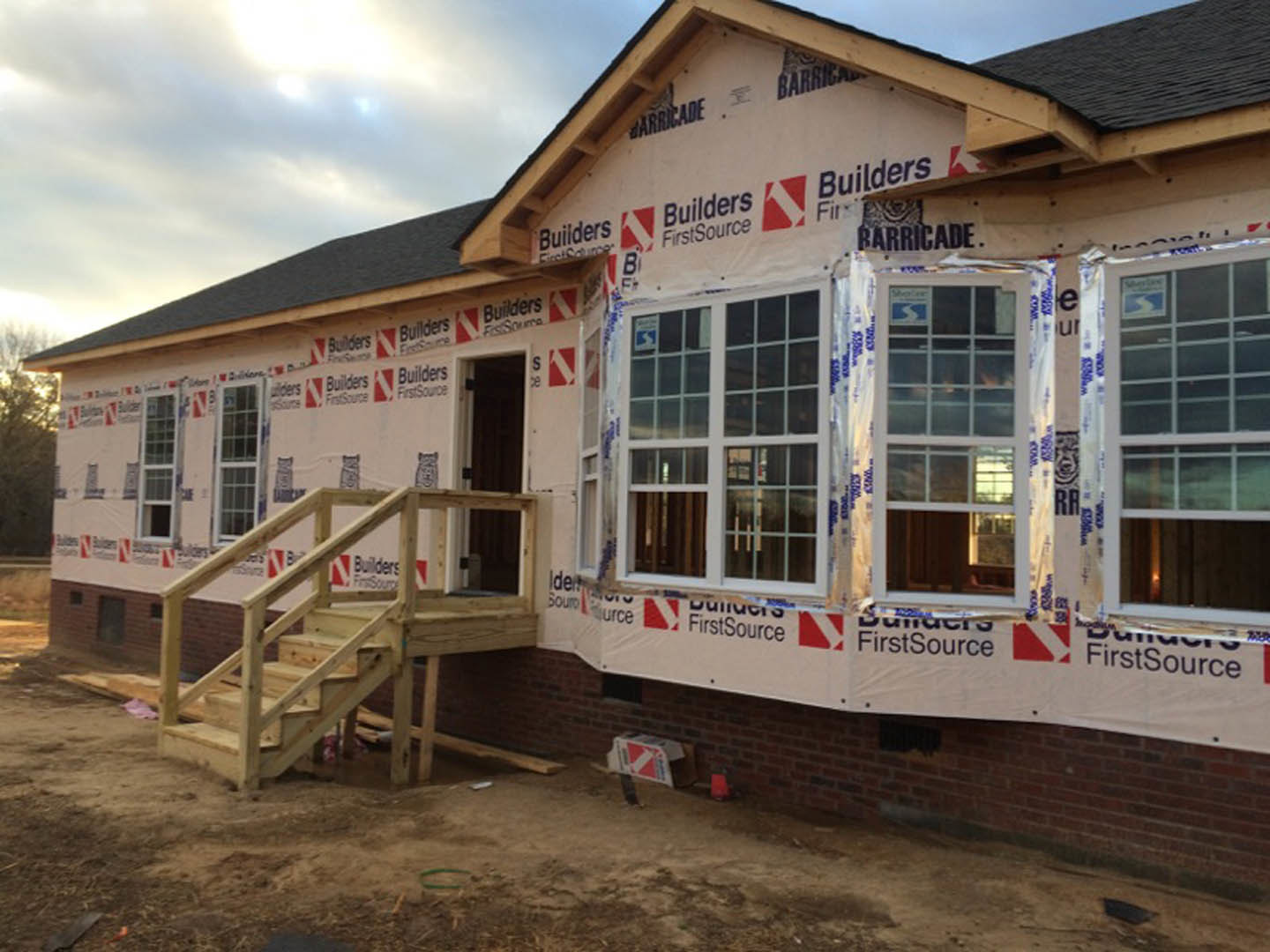 Two-story house under construction with exposed wooden staircase, multi-pane windows, unfinished exterior walls, and cloudy sky in background