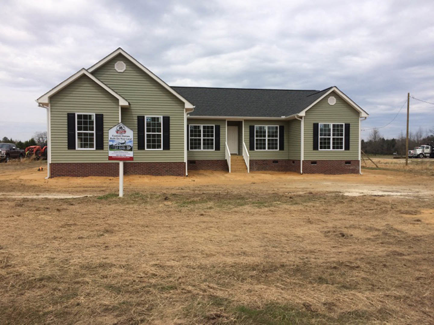 Green two-story house with white stairs and windows, brick retaining wall with a real estate sign, brown grass lawn, cloudy sky in background