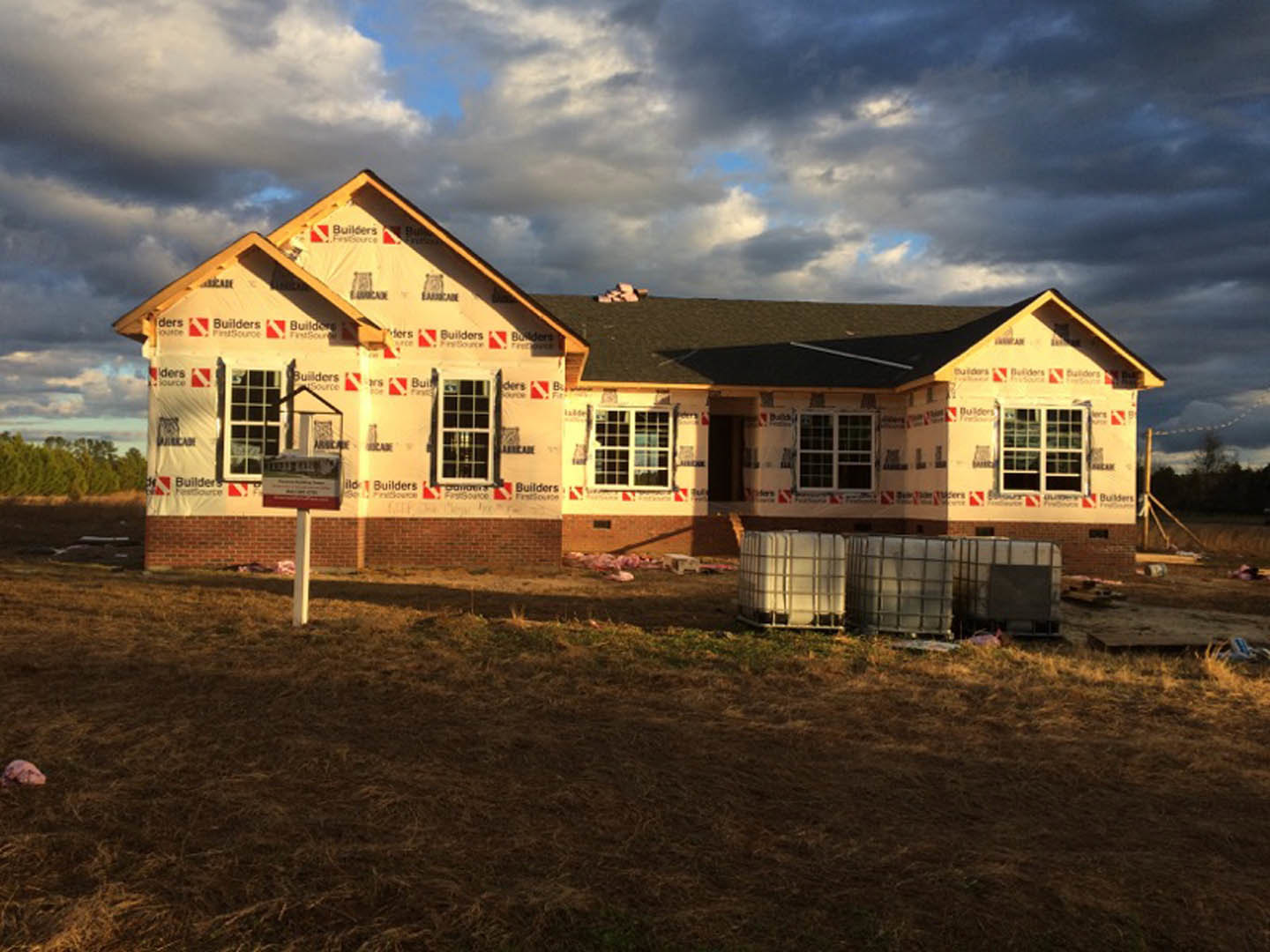 Two-story farmhouse with white siding, black-framed windows, and a grassy front yard, construction sign displayed near the entrance, cloudy sky overhead.