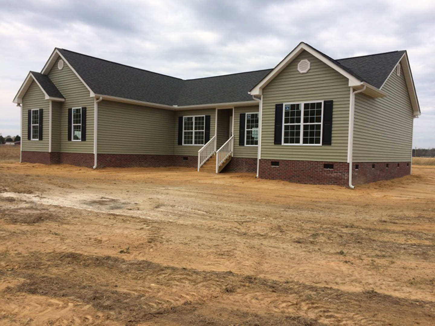 Two-story house with light siding, multi-pane windows, and white staircase railing, set beside a dirt road and bare ground under a cloudy sky.