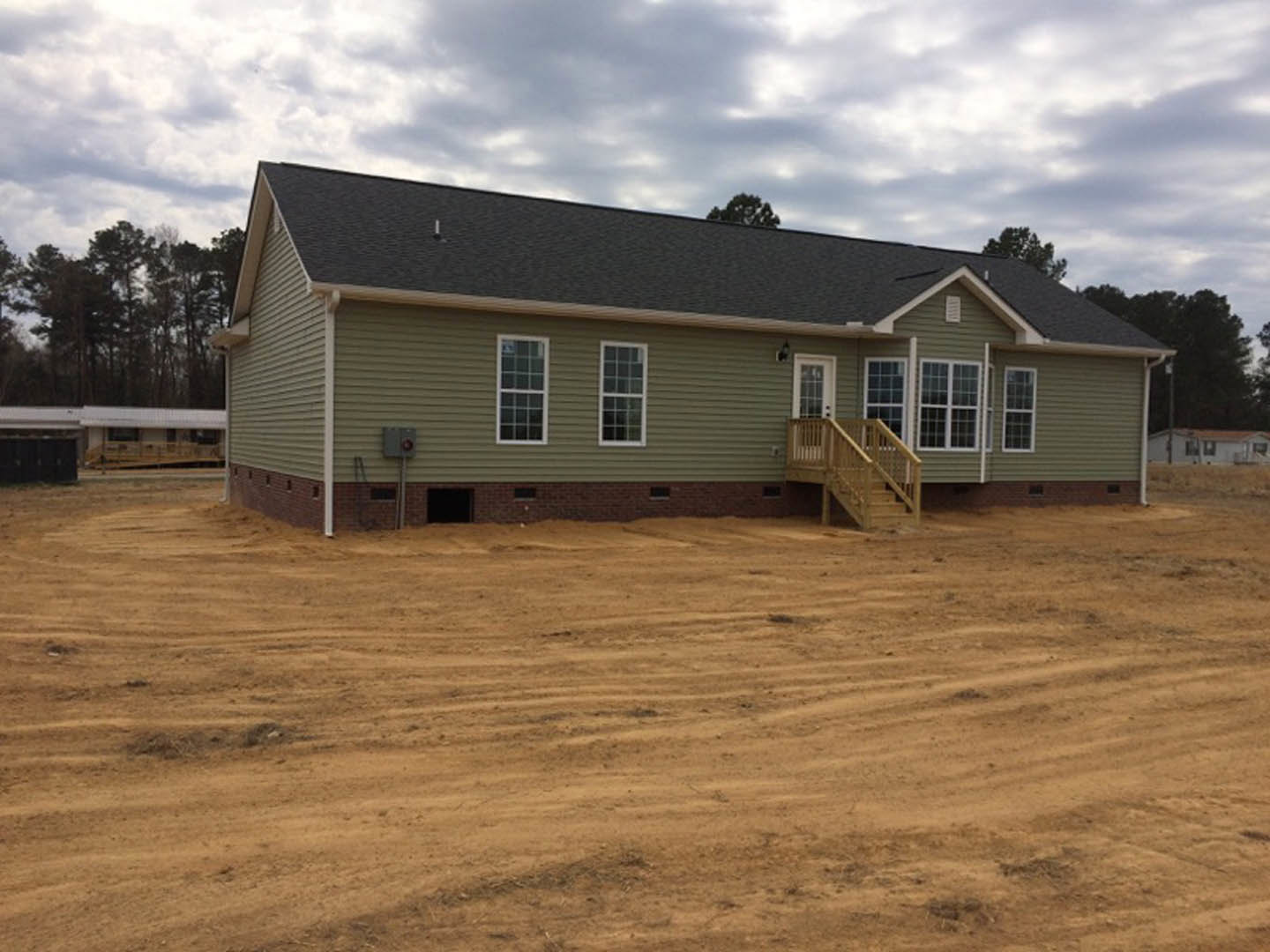 Wooden deck and staircase attached to partially constructed house, surrounded by dirt and mature trees, multi-pane windows visible on exterior.