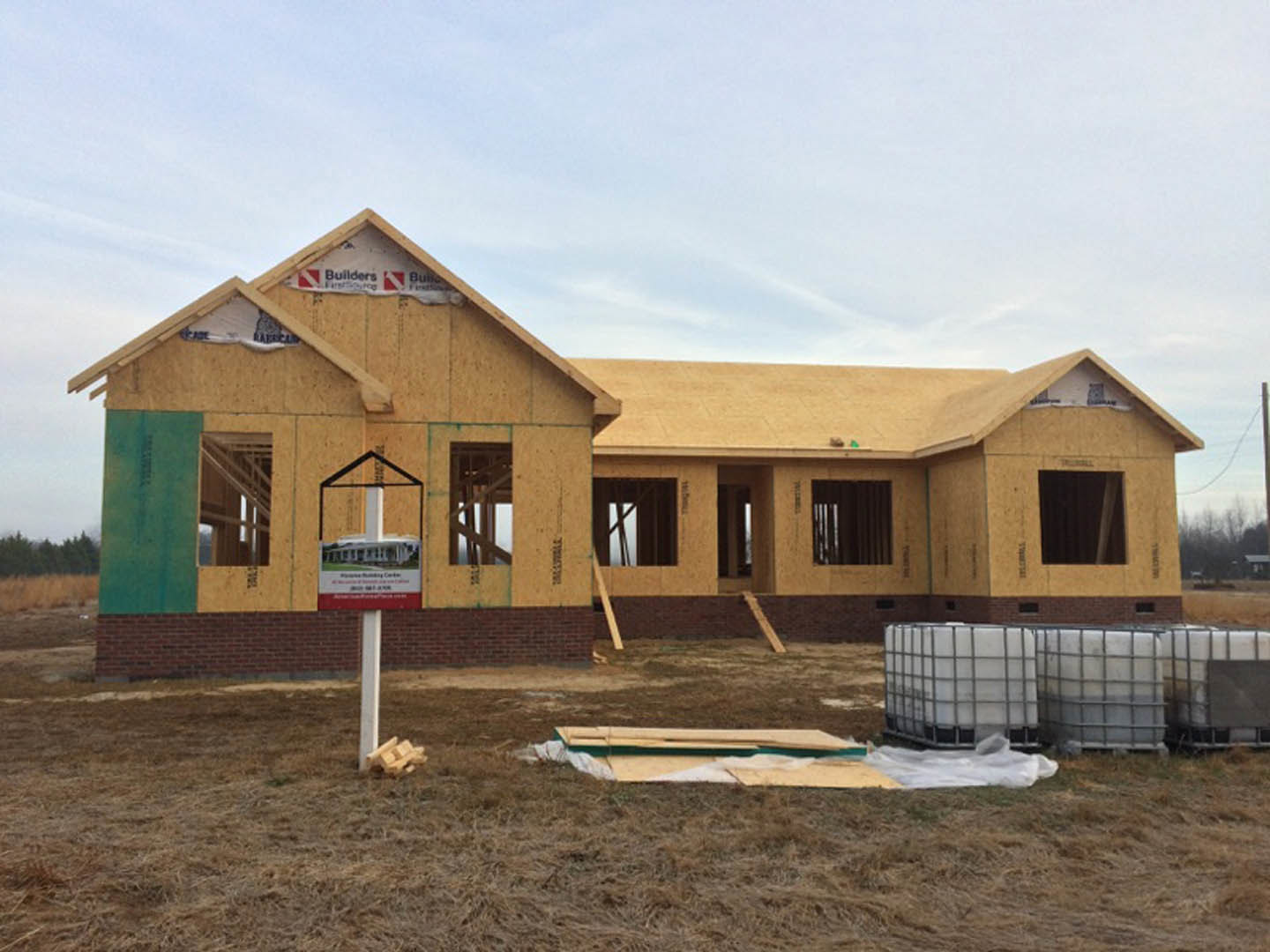 Brick house under construction with exposed framing, several installed windows, white plastic containers, wood pile, construction sign on brick wall, cloudy sky overhead