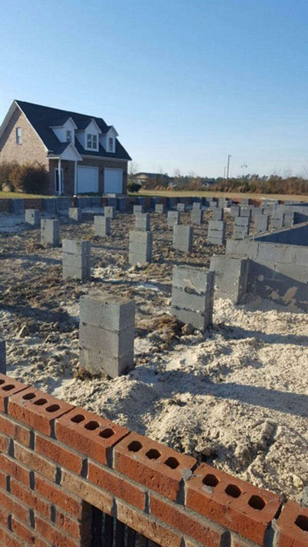 Two-story house with attached garage on a construction site, brick wall and concrete blocks in foreground, blue sky overhead
