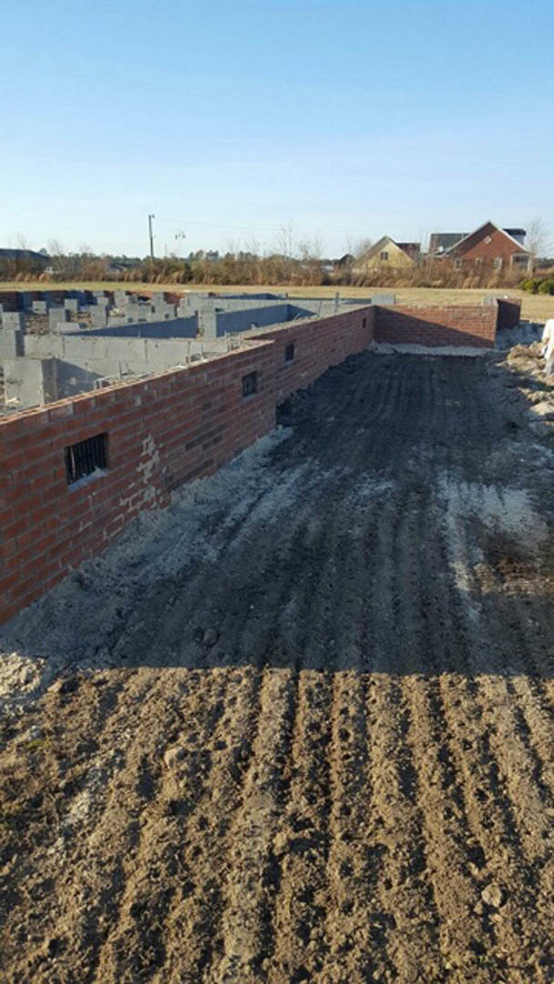 Red brick wall with small holes, metal-grated window, dirt ground alongside wall, blue sky with scattered clouds above