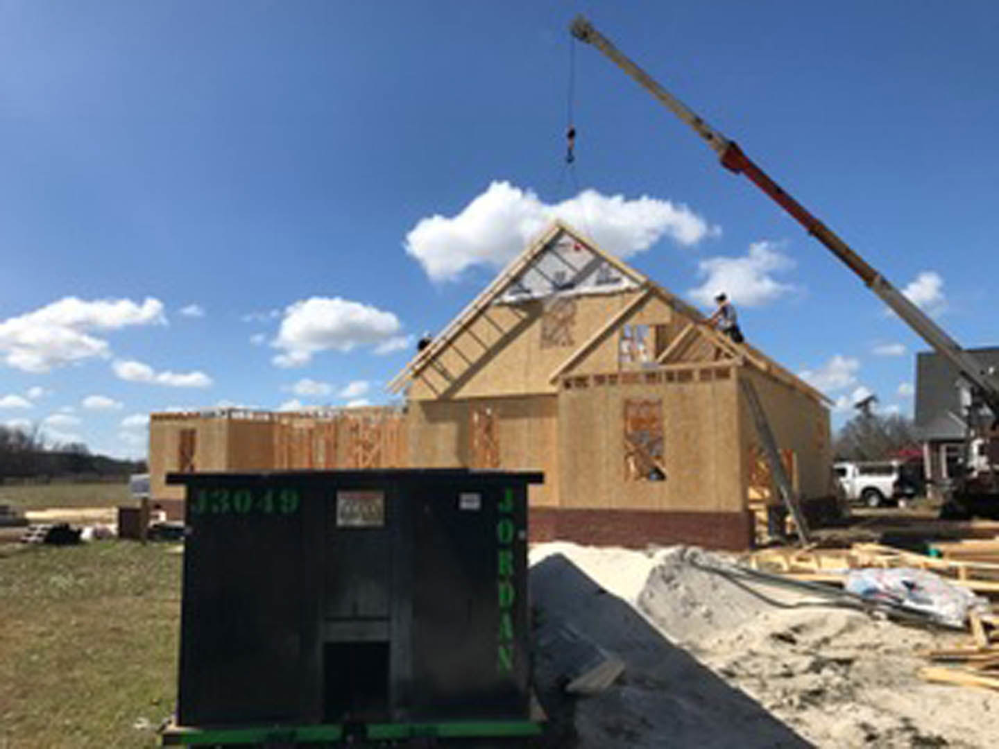 Crane hoisting modular black house with green doors above construction site, blue sky and clouds overhead, sand pile and pipe in foreground, trees and grass surrounding area