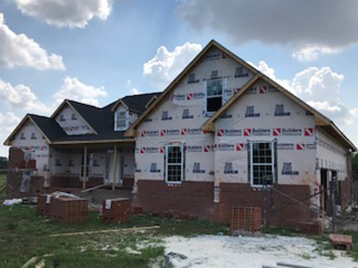 Framed house under construction with exposed plywood walls, grid-style window, and clear blue sky overhead