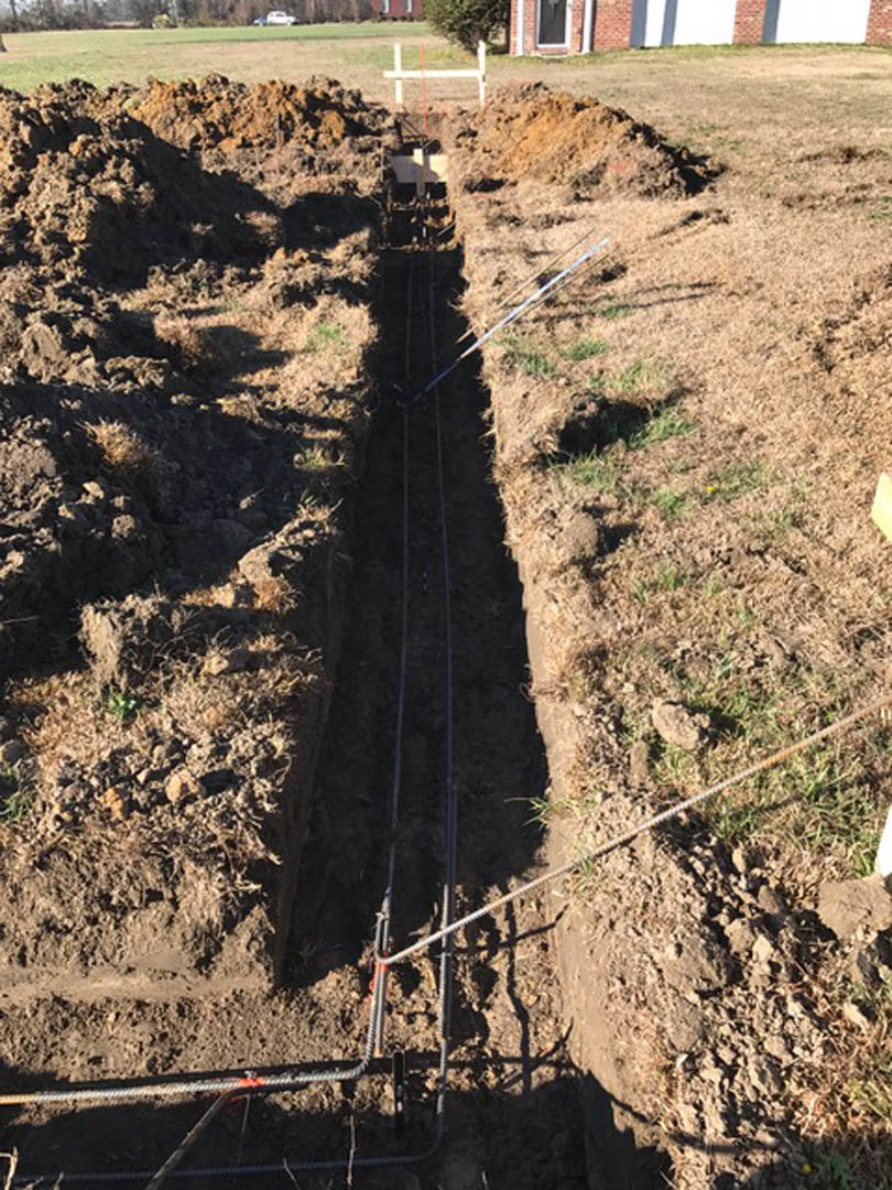 Open trench in soil with exposed electrical wires, surrounded by grass and small plants