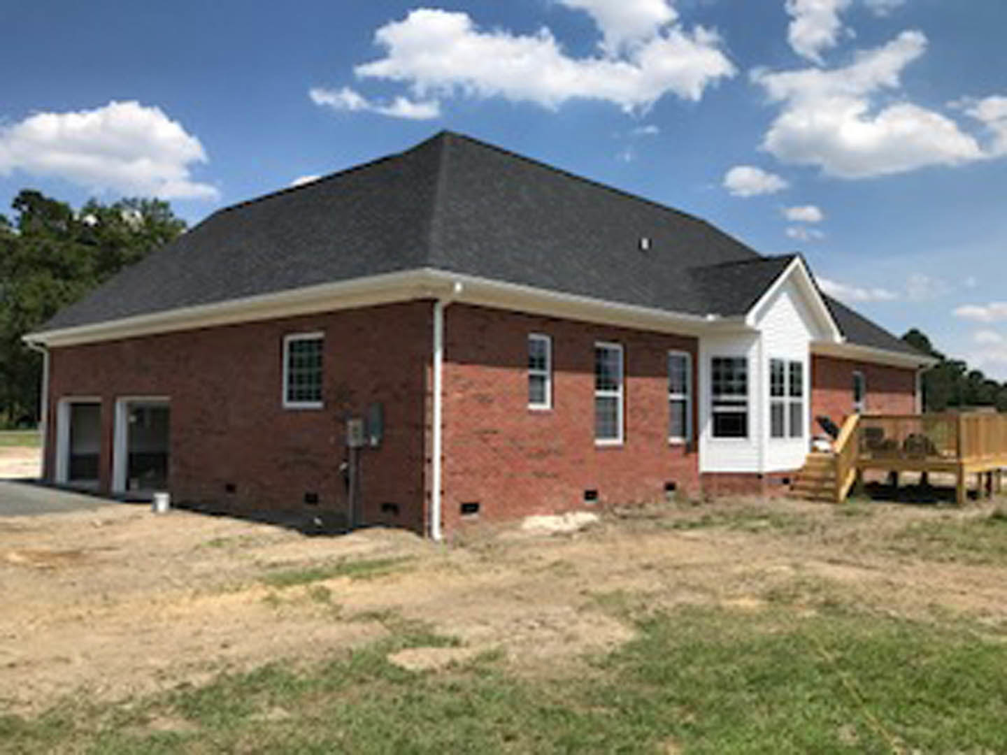 Red brick house with black roof, grid windows, grassy yard, and dirt patch under a partly cloudy blue sky