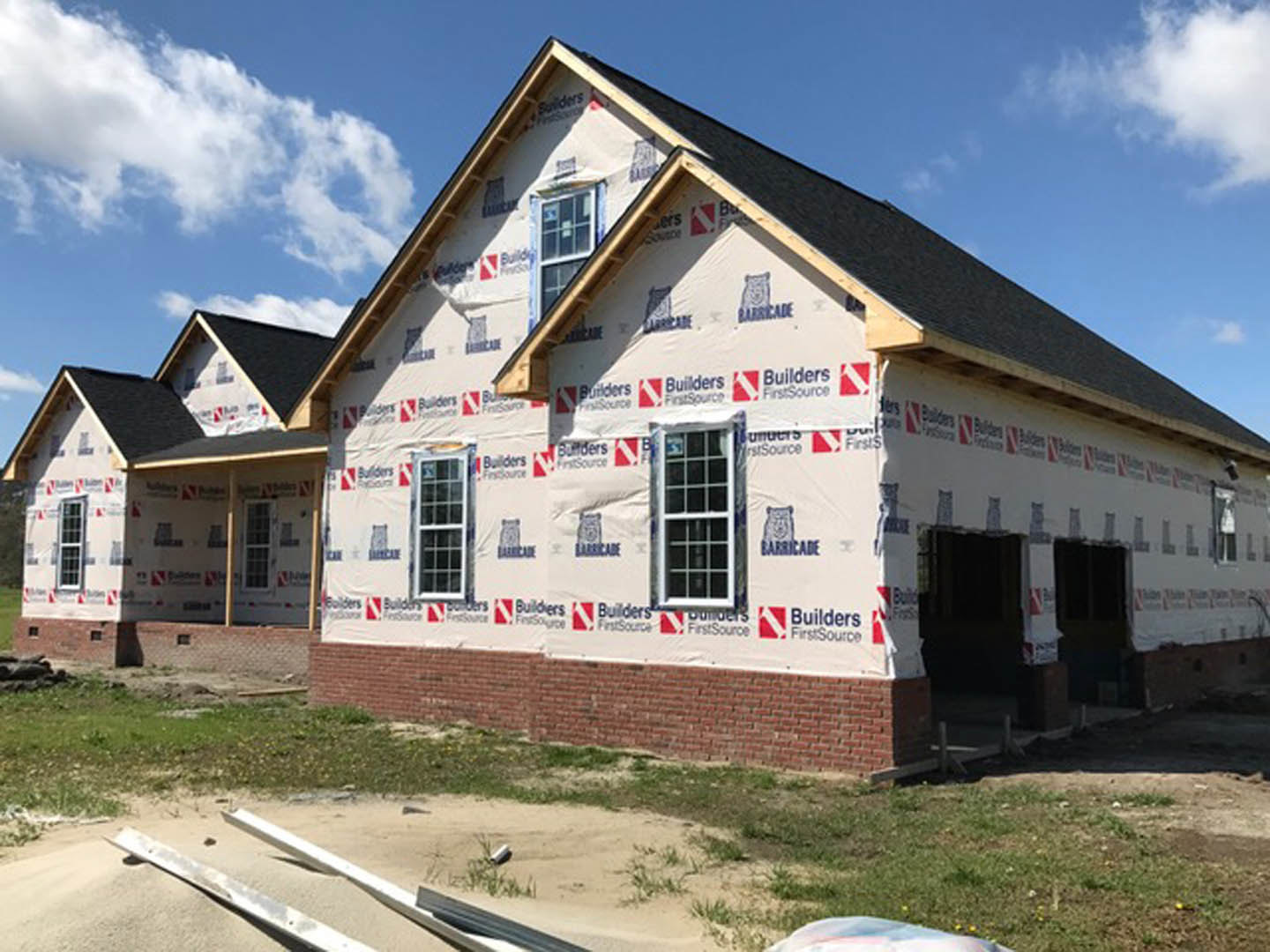 Partially built home with multi-pane window, plastic sheeting covering exterior walls, red and white bear logos on house wrap, dirt road in foreground, cloudy sky overhead