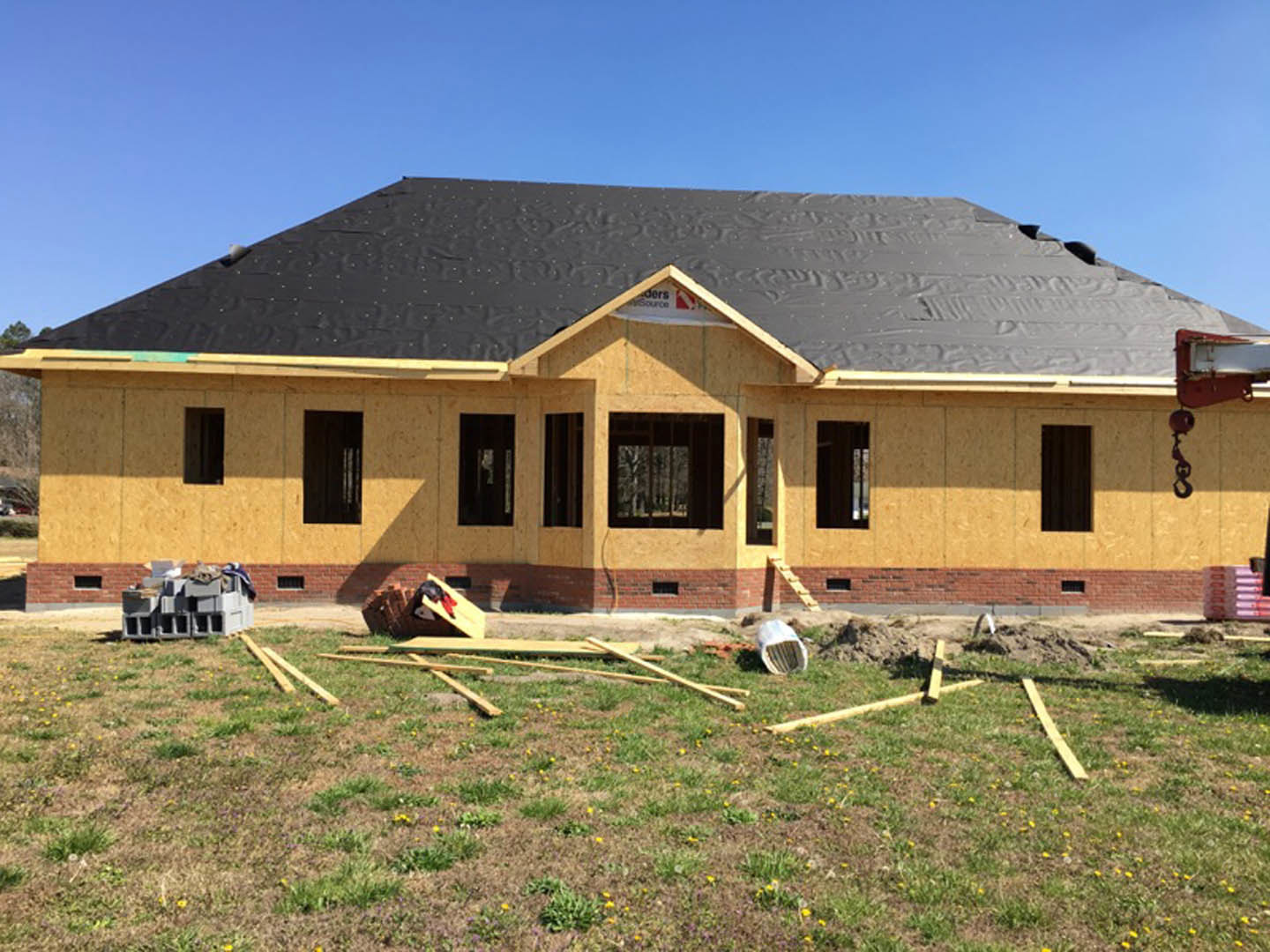 Partially built house with black roof, barred window, stack of grey blocks, and pile of wood on grassy yard