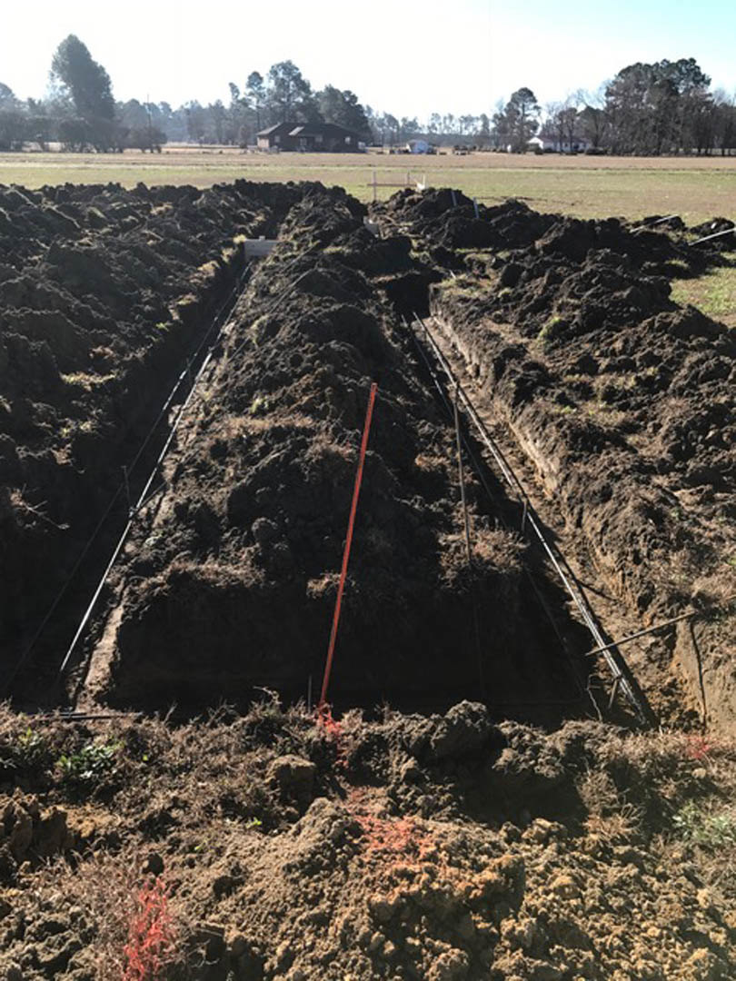 Dirt trench with exposed hole, metal pipes, red survey stake, surrounding soil and grass, blurred tree and plant in background