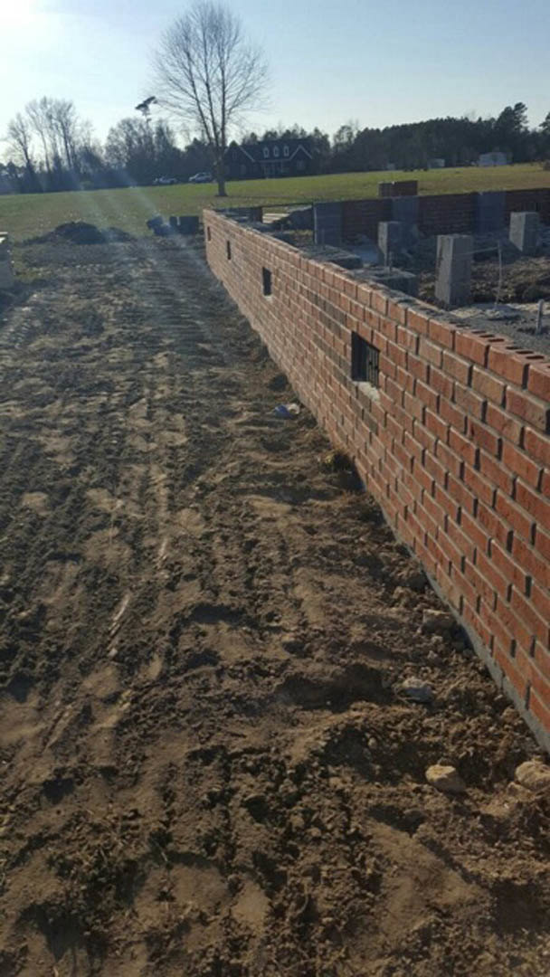 Red brick wall with a large hole, small windows, leafless tree nearby, dirt road and soil in foreground, metal fence and cloudy sky in background