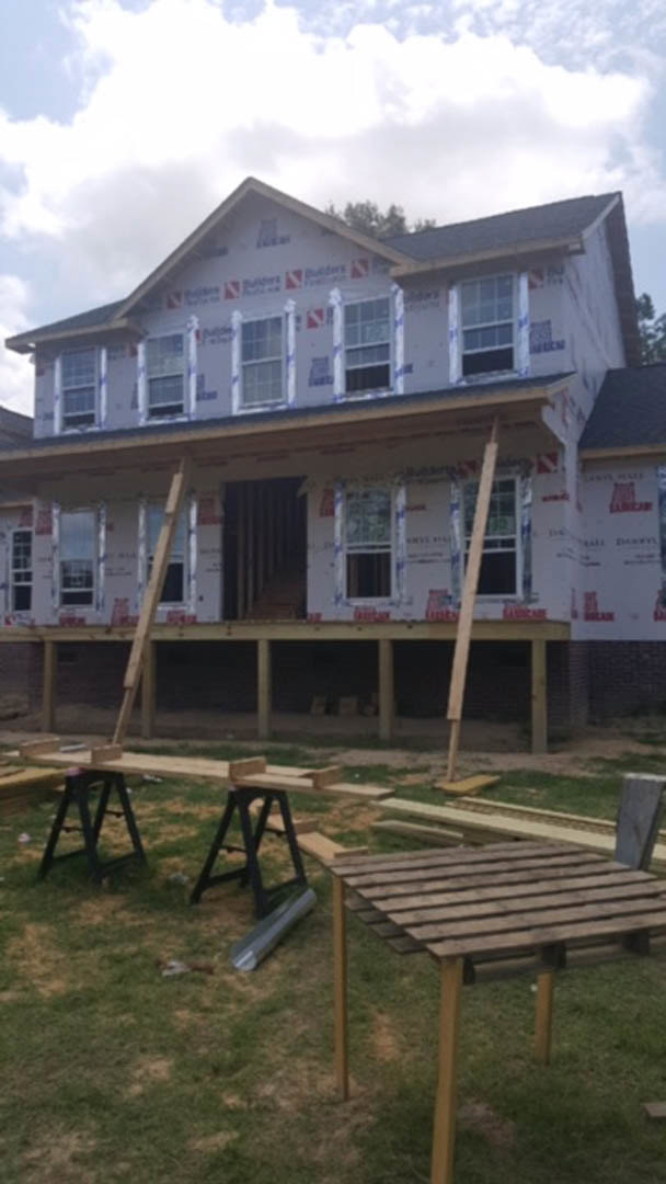 Wood-framed house under construction with exposed beams, stacked wooden planks on grass, metal pipe lying nearby, partially installed windows, and a covered porch area.