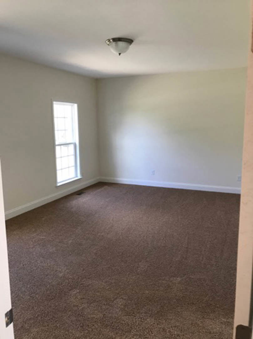 Bright room with white walls, brown carpet flooring, ceiling-mounted light fixture, and a window with a white frame