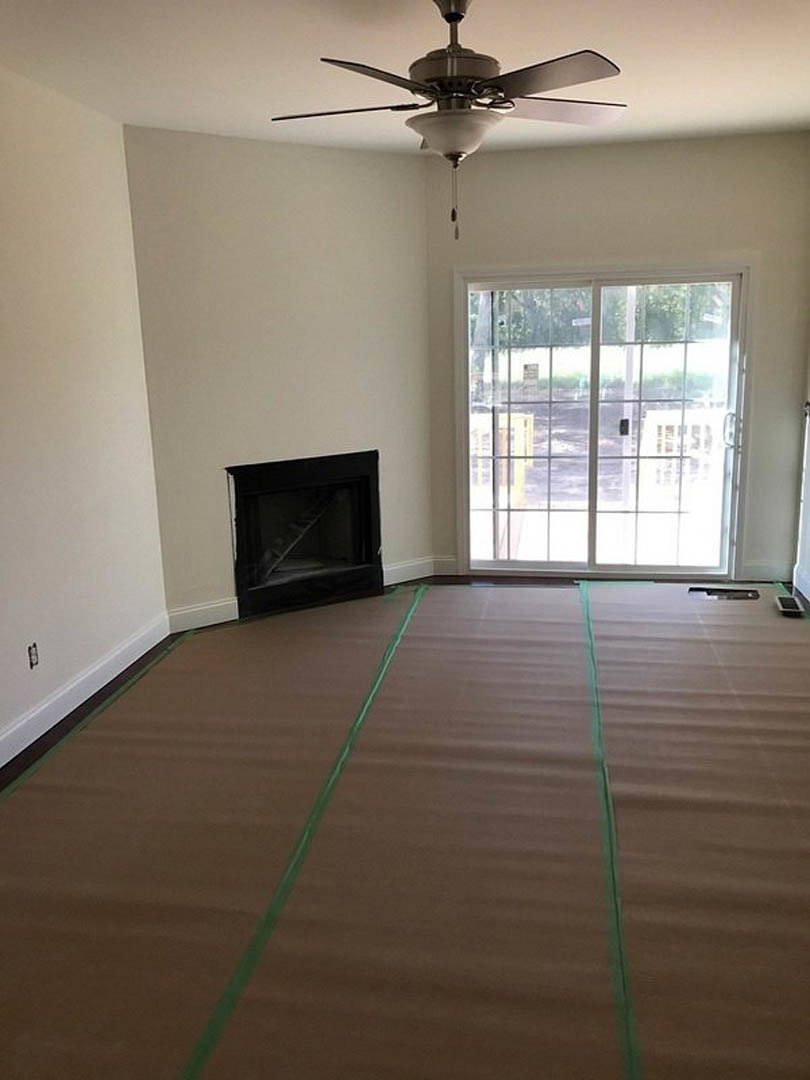 Living room with black fireplace, sliding glass door with white frame, brown floor marked with green tape, ceiling fan with light fixture, and staircase visible in background.