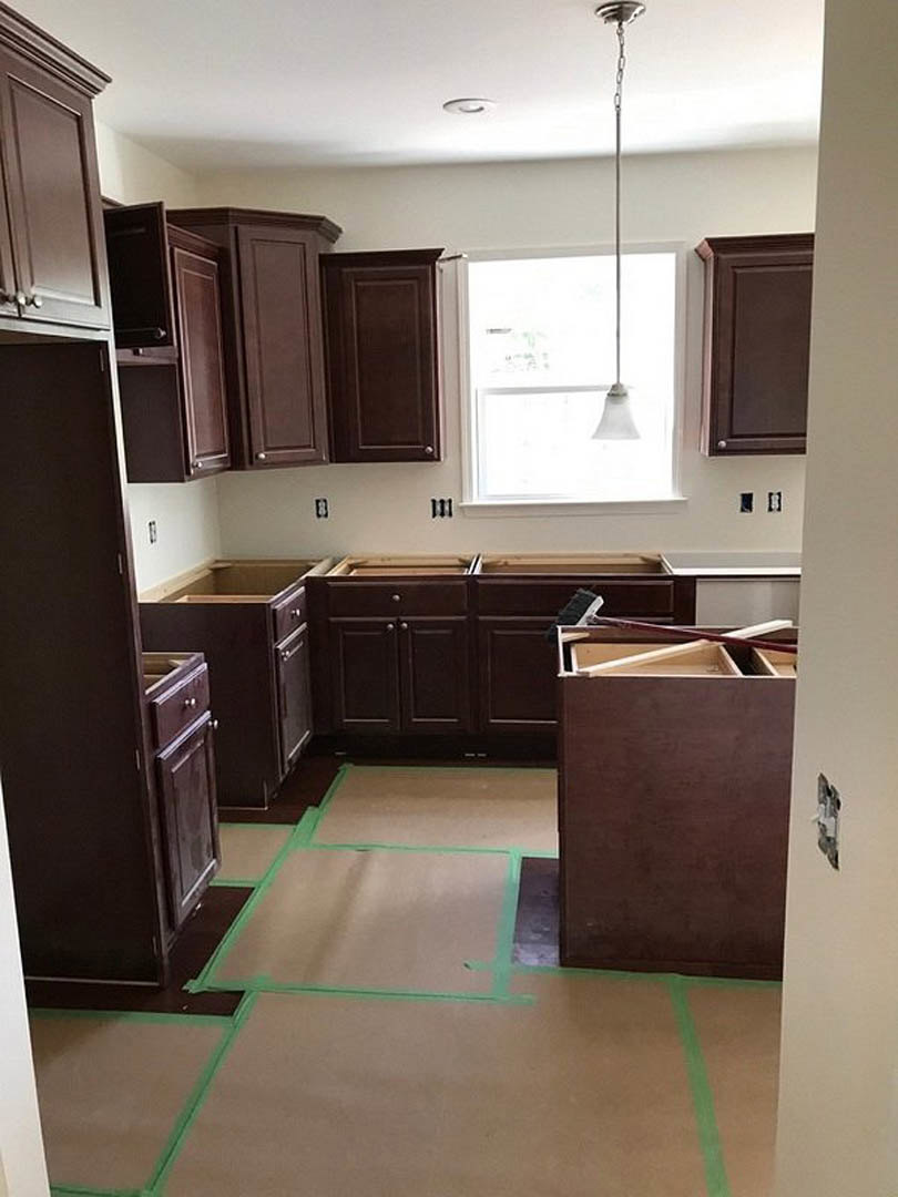 Kitchen with dark brown cabinets, black countertop, stainless steel sink, sunlight streaming through window, green tape on light-colored floor.