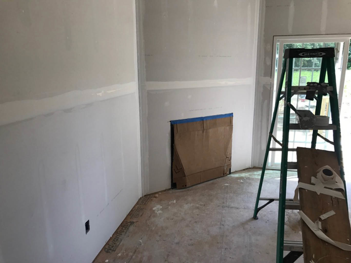 Aluminum ladder leaning against a white plaster wall with a strip of fresh paint, cardboard box nearby, roll of toilet paper on ladder, taped wood piece, dirty unfinished floor