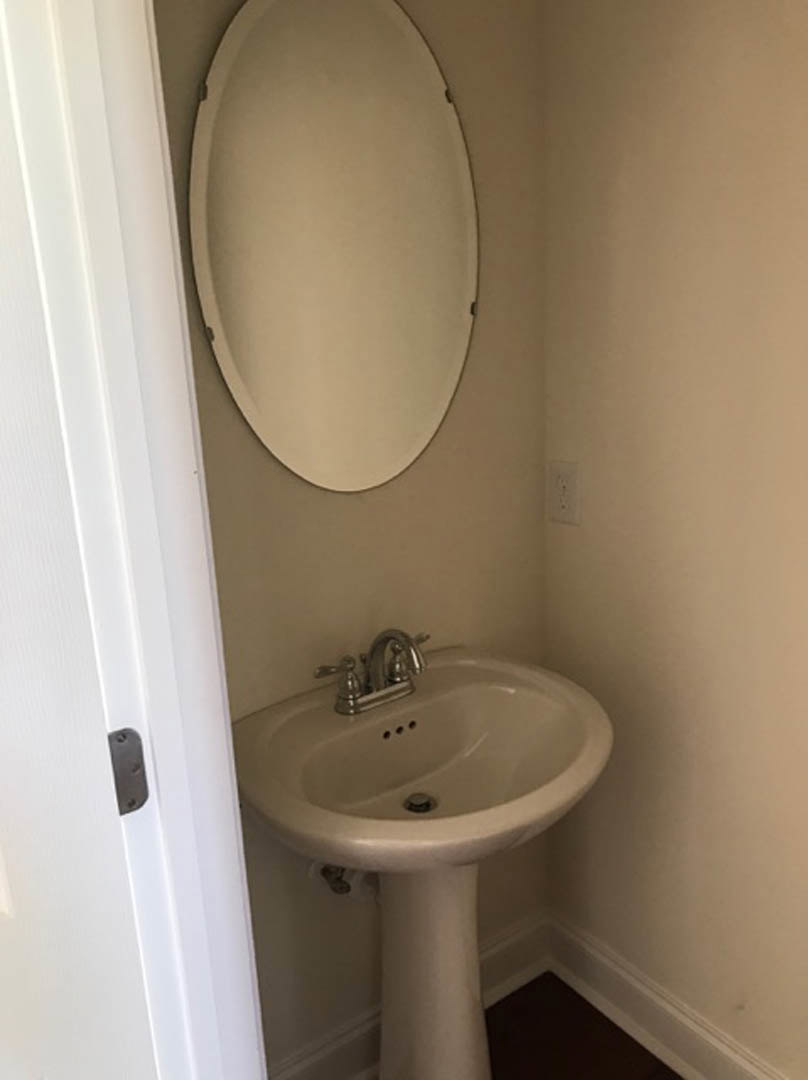 White ceramic sink with chrome faucet set into a light wood vanity, round mirror mounted above, gray tile backsplash and wall, bathroom accessories visible on countertop