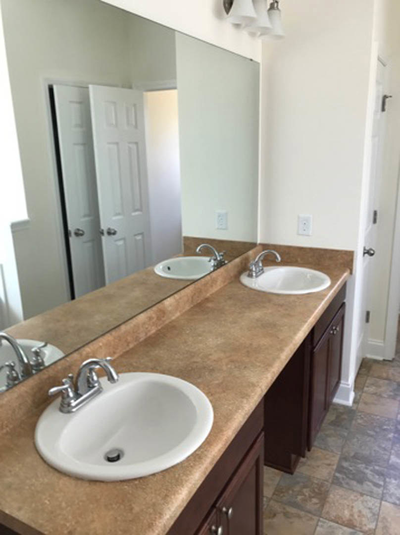 White double sinks with chrome faucets set in a stone countertop, large frameless mirror above, light gray tile backsplash, and modern bathroom accessories