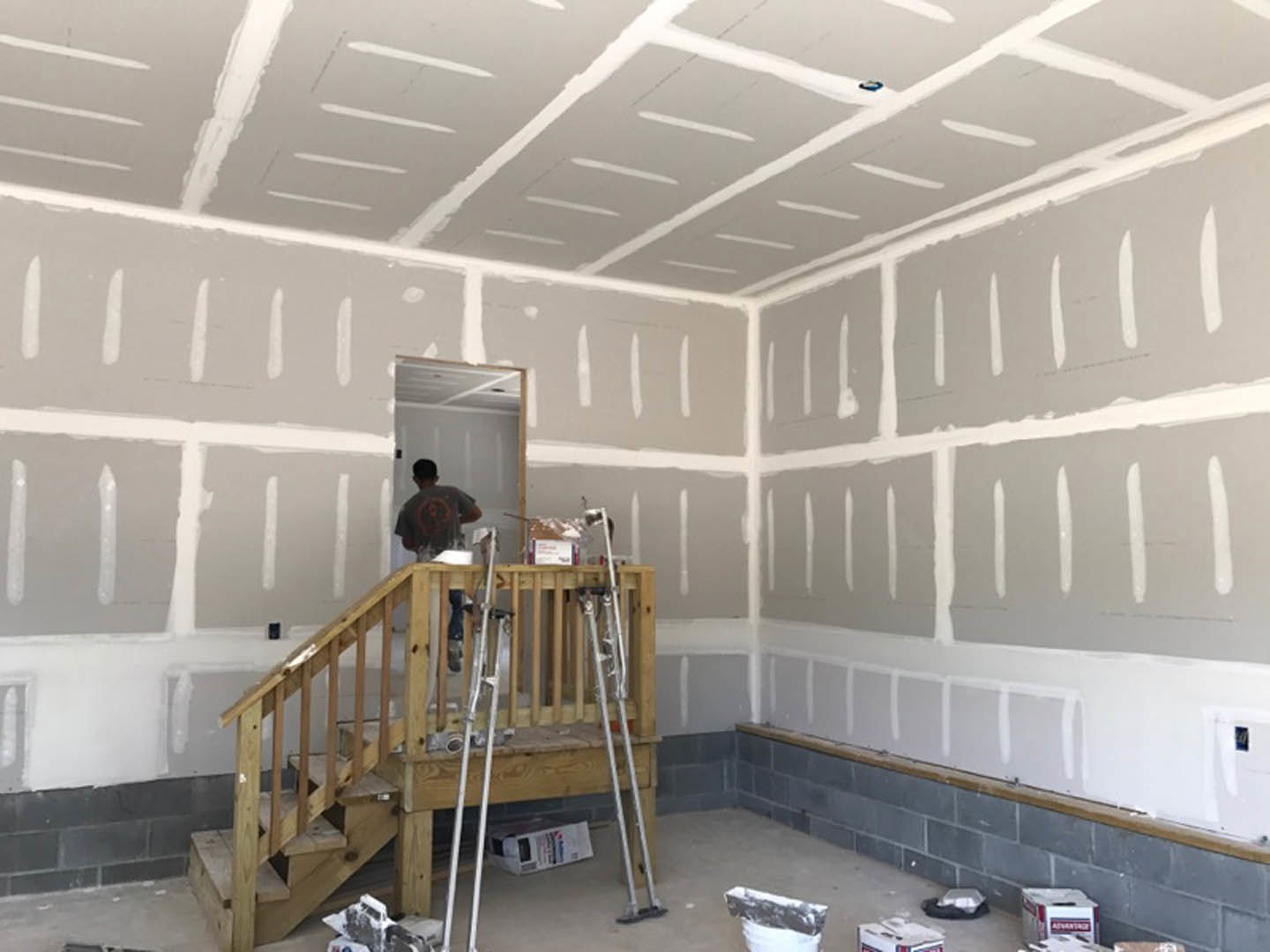 Man painting white wall while standing on staircase in room with plaster ceiling, wooden floor, and ladder nearby