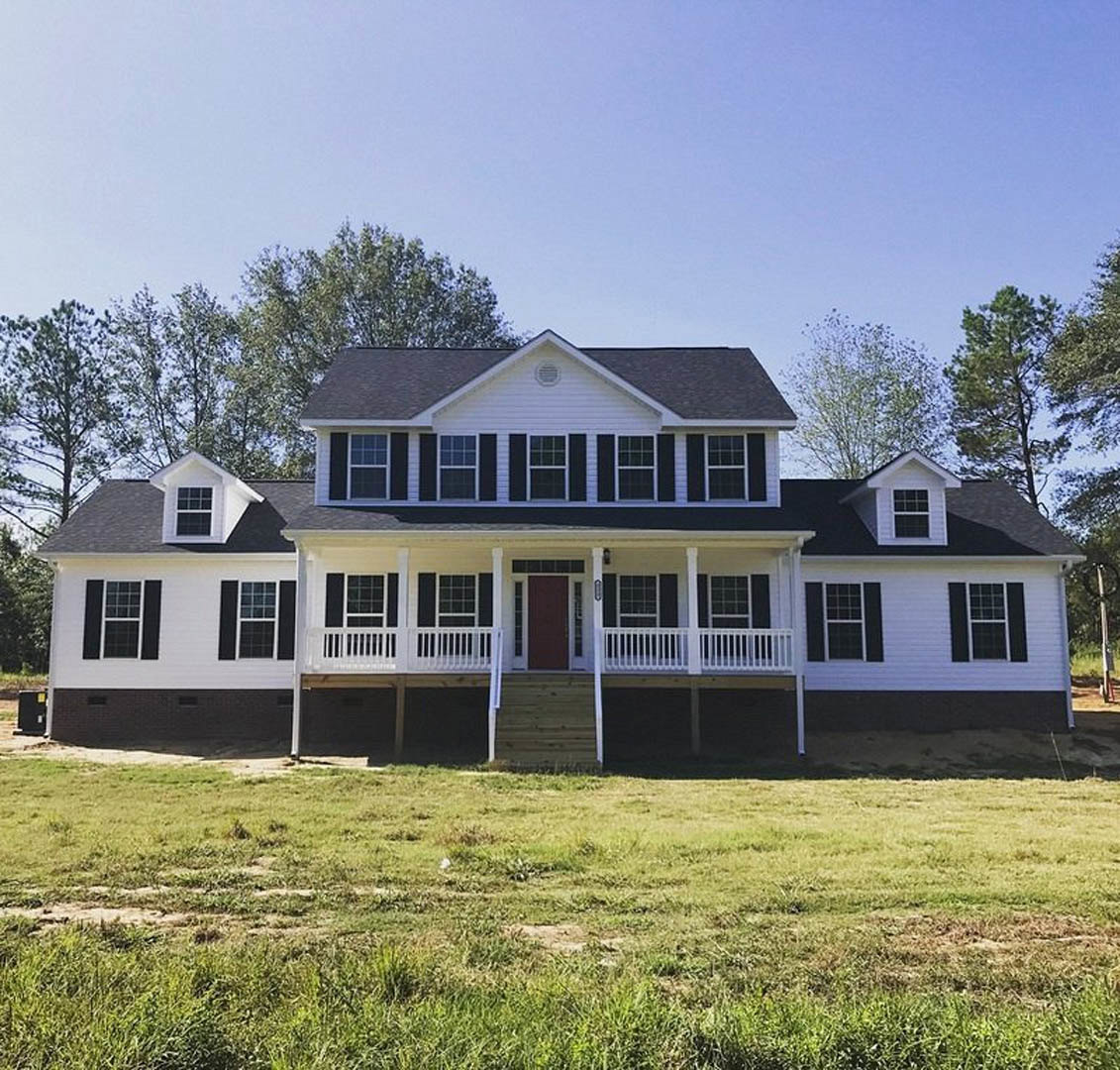White farmhouse-style home with red front door, covered porch, white-framed windows, and gabled roof vent, set on a grassy lawn with mature trees in the background