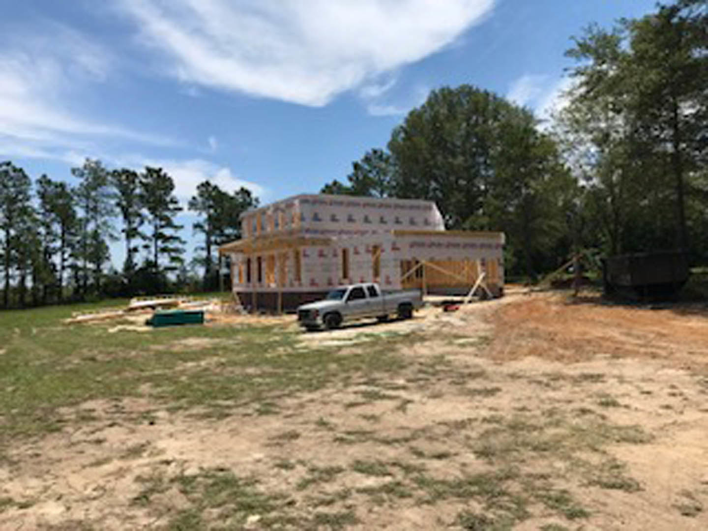 White truck parked on grassy lot in front of partially built house with exposed framing, surrounded by trees under blue sky with scattered clouds