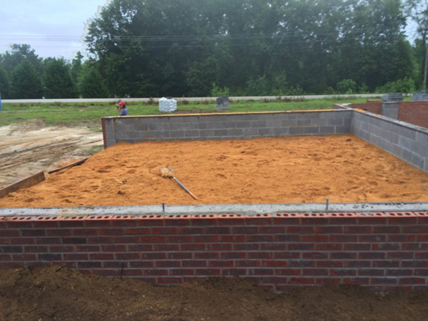 Brick foundation with exposed soil and scattered dirt, surrounded by outdoor trees and cloudy sky