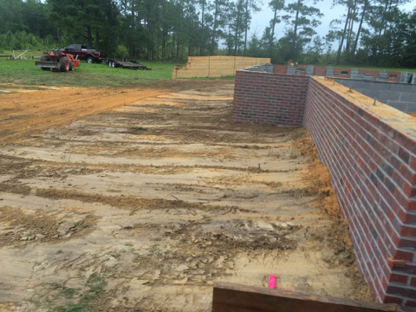 Dirt road bordered by a brick wall, tractor parked in the background, grassy patches and trees visible under a clear sky