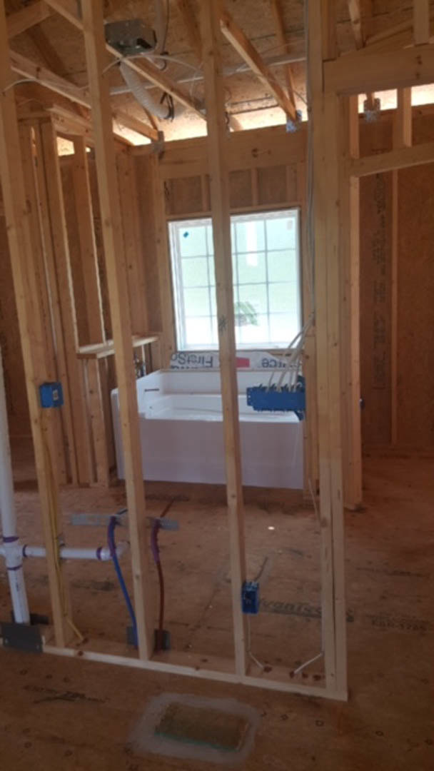 Bathroom under construction with exposed wood beams, unfinished walls, white bathtub, blue paint brush resting on tub edge, large window providing natural light