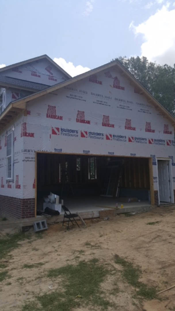 Framed house under construction with attached garage, exposed plywood walls, construction materials scattered on dirt ground, black folding chair near entry, large windows