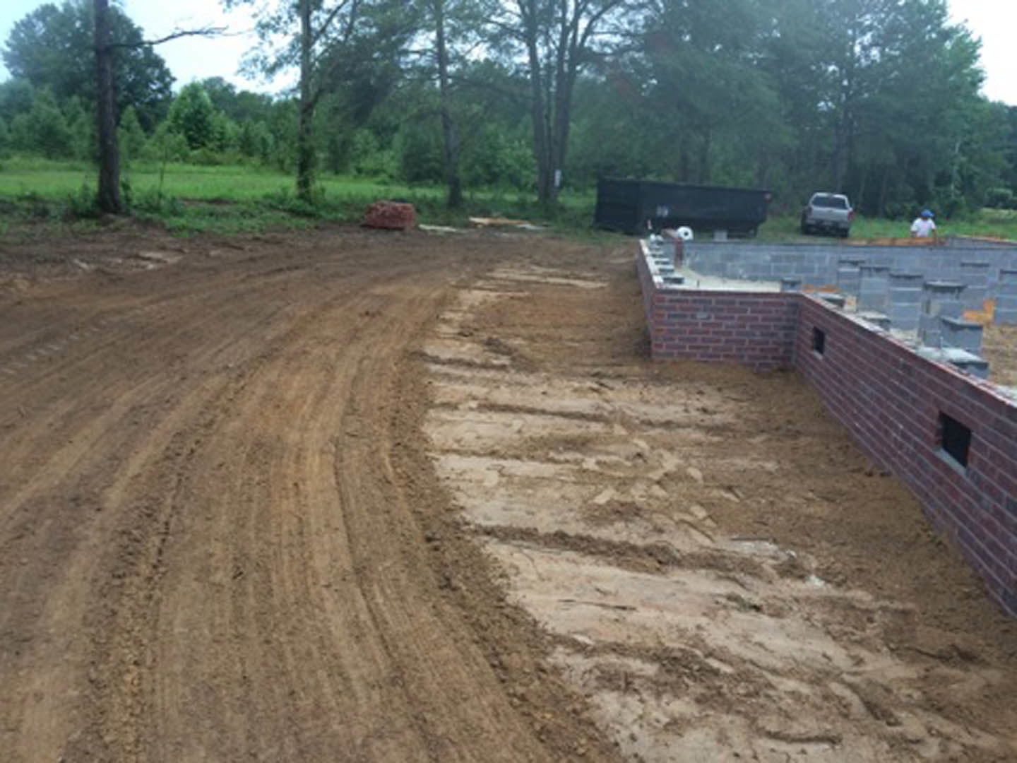 Dirt road bordered by a brick wall with a hole, surrounded by grass and trees under a clear sky