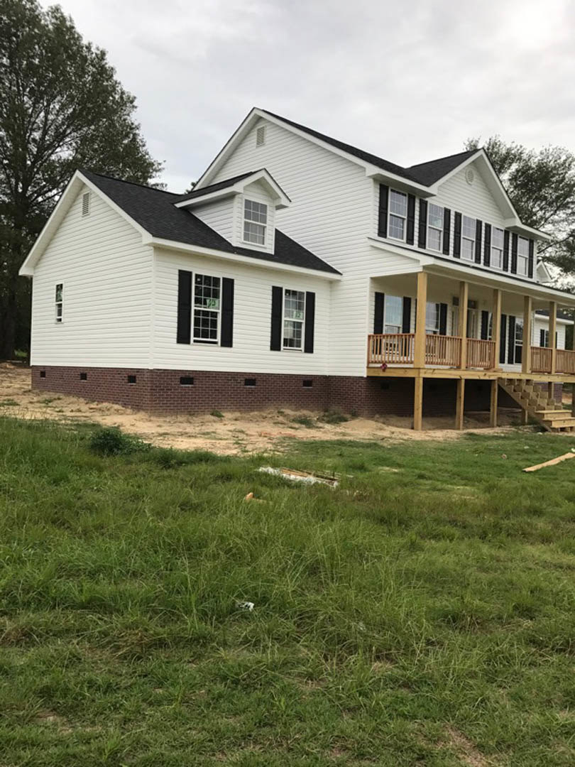 White siding house with covered porch, green lawn, large windows, and tree in the background under partly cloudy sky